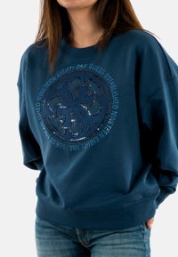 Sudadera de color azul marino con un logo redondo de lentejuelas y texto en azul claro. Presenta un amplio escote de barco y un corte holgado. Tejido texturizado.