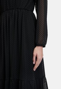 Schwarzes Kleid mit Polka-Dots, transparenten langen Ärmeln, gerafft in der Taille und mit gestuftem Saum. Leichte Textur und subtiler Glanz steigern die visuelle Anziehung.