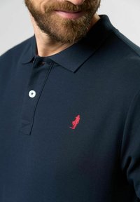 Marineblå polo-shirt i tekstureret stof med krave med tre knapper og et lille rødt broderet logo på venstre bryst. Hvid knap synlig.