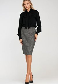 Kobieta w czarnej bluzce z długim rękawem i kokardą z przodu, w szarej spódnicy ołówkowej i czarnych szpilkach, stojąca na tle jednolitego jasnego tła.