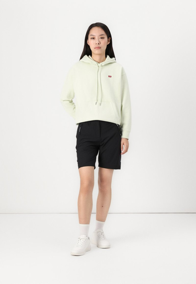 Felpa verde chiaro con cappuccio e logo, abbinata a pantaloni corti neri. Il modello indossa scarpe da ginnastica bianche e calzini alla caviglia. Design semplice, abbigliamento casual.