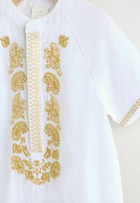 Vêtement blanc avec des broderies dorées complexes sur le devant et les manches. Tissu doux avec une texture lisse et un design sans col.
