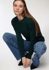 Ciemnozielony sweter dzianinowy z okrągłym dekoltem, w zestawieniu z jasnoniebieskimi spodniami o szerokich nogawkach. Modelka jest w pozycji klęczącej, ma na sobie czarne, spiczaste buty.