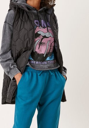 Personne portant une veste sans manches matelassée noire, un sweat à capuche gris délavé avec un motif de langue graphique, et un pantalon de survêtement bleu à cordon, les mains dans les poches.