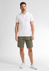 Bílý polo tričko s malými, barevnými vzory, spojené s olivově zelenými cargo šortkami. Model má na sobě bílé tenisky. Jednoduché pozadí.