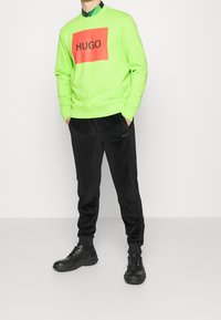 Felpa verde neon con un grande rettangolo rosso che presenta il testo "HUGO"; abbinata a pantaloni in velluto nero; scarpe nere con suole testurizzate.