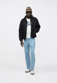 Schwarze Daunenjacke mit hohem Kragen, weißes Graphic-T-Shirt, hellblaue Jeans und eine mehrfarbige gestreifte Strickmütze. Legere Sneakers.