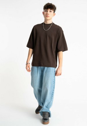 Młody mężczyzna idący, ubrany w luźną ciemnobrązową koszulkę, jasnoniebieskie obszerne dżinsy, czarne tenisówki, srebrny łańcuch na szyi i bransoletkę na białym tle.