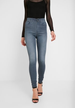 Kobieta ubrana w dopasowane niebieskie dżinsy z wysokim stanem, przezroczystą czarną bluzkę z długim rękawem oraz czarne sandały na obcasie z odkrytymi palcami, stojąca na jednolitym tle.