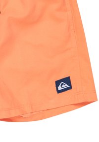 Oranžové plavkové šortky z ľahkej tkaniny s hladkou štruktúrou, so značkovou nášivkou v námorníckej modrej farbe na spodnej časti.