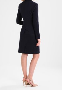 Kvinna iklädd en tight svart kavaj och kjol med beige högklackade skor, stående med ryggen vänd mot en vit bakgrund.