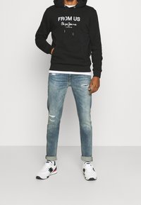 Felpa nera con testo bianco, jeans in denim con dettagli sfilacciati e sneakers nere, bianche e rosse. Outfit casual con un look a strati.