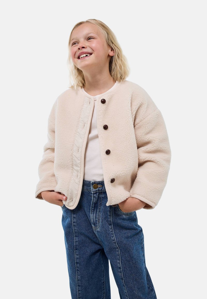 Veste polaire beige avec encolure ronde, fermeture à boutons et poches latérales, portée sur un haut blanc et un jean bleu, avec une coupe décontractée.