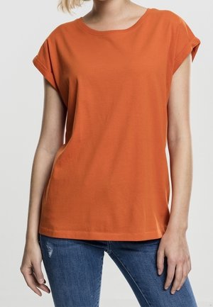 Woman wearing a plain orange short-sleeve T-shirt and blue jeans, standing with relaxed posture against a neutral background.