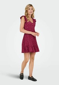 Burgundy klänning med vita prickar, volangärmar, V-ringning och utställd fåll. Tyget verkar vara lätt och texturerat.