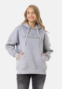 Grauer Kapuzenpullover mit Kordelzugkapuze, Fronttaschen und gesticktem Logo. Weicher Stoff und lockere Passform, kombiniert mit dunklen Jeans.