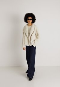 Λευκό φουντωτό μπουφάν με γιακά φουλάρι, λεπτομέρεια από κουμπιά, συνδυασμένο με σκούρα φαρδιά τζιν. Το μοντέλο φοράει γυαλιά ηλίου και ένα ανοιχτόχρωμο τοπ.