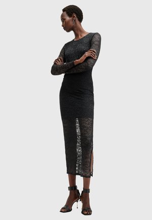 Zwarte kanten jurk met lange mouwen en een aansluitend silhouet, voorzien van doorschijnende panelen en zijsplitten. Gecombineerd met zwarte open teen hakken.