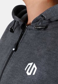 Sivá mikina s kapucňou na zips, so šnúrkou na stiahnutie a bielym logom na hrudi. Má jemnú textúru a tesný strih.