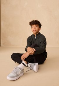 Maglione grigio a costine con collo con zip, pantaloni neri, sneakers Nike grigie con suole bianche e calzini bianchi con strisce nere.