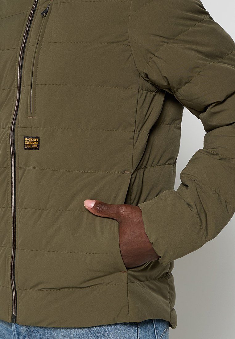 Mano derecha en el bolsillo de una chaqueta acolchada verde oliva con cremallera y una pequeña etiqueta negra y amarilla de G-Star en el pecho.