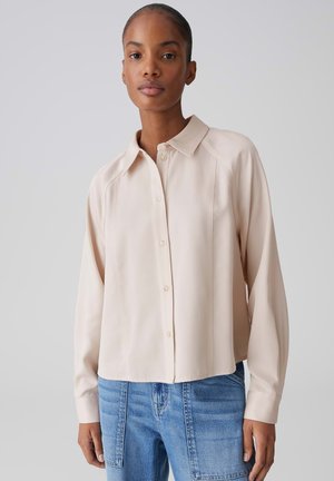 Vrouw die een lichtbeige blouse met knopen draagt, voorzien van een puntige kraag en lange mouwen, gecombineerd met high-waisted blauwe spijkerbroek.
