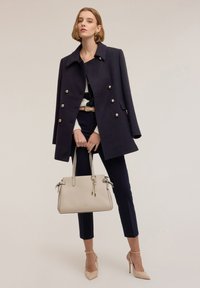 Granatowy dwurzędowy płaszcz z złotymi guzikami, biały sweter, dopasowane granatowe spodnie, beżowa torebka i beżowe buty na wysokim obcasie.