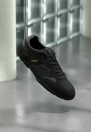 Black athletic sneaker with "HUGO" logo in gold on the side, black laces, and textured panels, floating above a polished concrete floor.