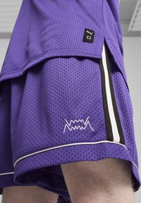 Pantalones cortos deportivos morados de tela de malla, con una franja lateral blanca y negra, superficie texturizada y un distintivo logotipo bordado.