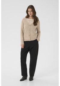 Beige cardigan with button closure, V-neck, lace detail underneath, paired with black tapered trousers and ankle boots. Smooth texture, casual fit.