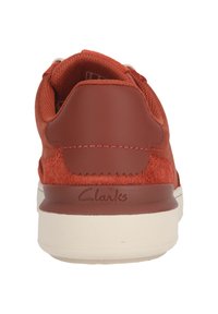 Punaiset samettitopaiset ja sileät nahkakengät, joissa on pehmeä kaulus, kermaisen värinen ulkopohja ja kohokuvioitu "Clarks"-logo kannassa.