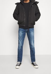 Man i svart hujacka, svart sweatshirt, blå jeans och svarta höga sneakers som står mot en enfärgad bakgrund.