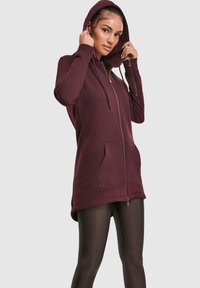 Temno rjavi pulover s kapuco na zadrgo, z žepi spredaj in dolgimi rokavi, s gladko teksturo in zlatimi detajli na zadrgi.