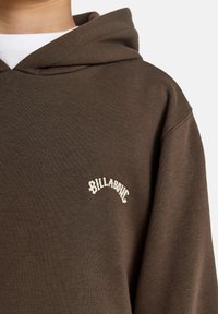 Brun hettegenser med en hvit brodert "Billabong"-logo nær halsen, med avslappet passform og snorhette.