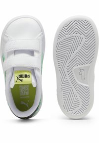 Des chaussures Puma blanches avec une tige en cuir lisse, des accents verts, deux bandes Velcro et une semelle en caoutchouc texturé avec un motif anti-marking.