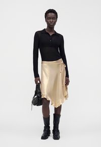 Donna con top nero a maniche lunghe, gonna avvolgente dorata lucida con orlo arricciato, stivali neri a metà polpaccio, che tiene una borsa nera.