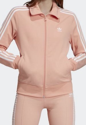Osoba ubrana w brzoskwiniowy dres Adidas z białymi paskami na rękawach i spodniach, ręce w kieszeniach kurtki, stojąca na jednolitym tle.