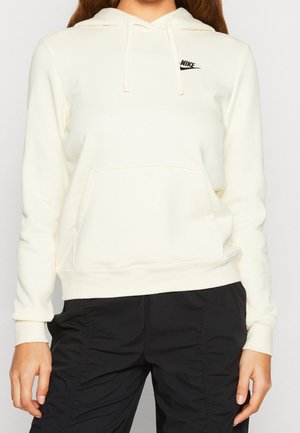 Femme portant un sweat à capuche Nike blanc avec poche à l’avant et un pantalon noir, debout devant un fond clair uni.