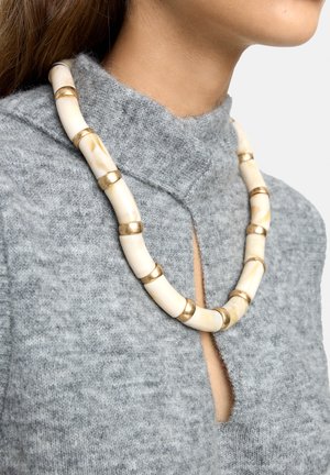 Naszyjnik z koralików wykonany z kremowego materiału ze złotymi akcentami, o zaokrąglonym, segmentowym wzorze i gładkiej fakturze, noszony na szarym topie.