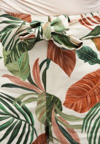 Κοντινή λήψη δεμένης ζώνης από ύφασμα σε παντελόνι με τροπικά πράσινα και σκουροκόκκινα φύλλα πάνω σε ανοιχτό φόντο.