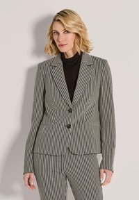 Frau mit blonden Haaren, die ein schwarzes Oberteil unter einem schwarz-weißen Hahnentrittmuster-Blazer und passenden Hosen trägt, steht vor einer hellen Wand.