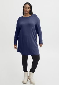 Abito tunica oversize blu navy con maniche lunghe, texture morbida, scollatura rotonda e spacchi laterali, abbinato a leggings neri e stivali alla caviglia color crema.