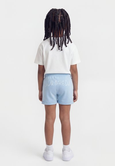 Παιδί με στριφτά πλεξούδες, που φοράει λευκό μπλουζάκι, ανοιχτό μπλε σορτς με τη σήμανση «DSQUARED2», λευκές κάλτσες και λευκά αθλητικά παπούτσια, όρθιο με την πλάτη στο θεατή.