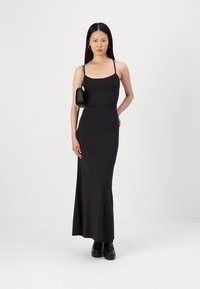 Črna, oprijeta, brezrokavna maxi obleka z okroglim izrezom, gladko teksturo in minimalističnim dizajnom, kombinirana z majhno črno torbico.