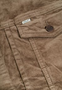 Tela de pana marrón con una superficie texturizada, que presenta un bolsillo de parche y un acento de botón de marca. Etiqueta blanca en el borde superior.