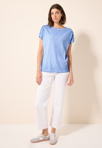 Top azzurro chiaro a maniche corte con vestibilità rilassata e maniche arrotolate, abbinato a pantaloni bianchi a gamba larga e sneakers grigie. Texture liscia.