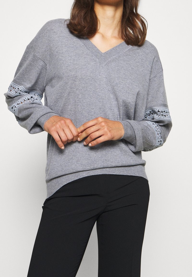 Femme portant un pull gris à col en V avec des détails en dentelle sur les manches et un pantalon noir, debout devant un fond uni.