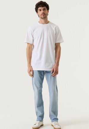 Mann steht und trägt ein einfaches weißes T-Shirt mit kurzen Ärmeln, hellblaue Jeans und weiße Turnschuhe vor neutralem Hintergrund.