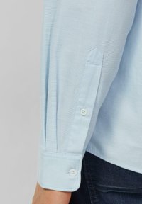 Chemise bleu clair à manches longues avec un poignet à boutons, présentant une texture lisse et une coupe ajustée avec un léger éclat.