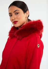 Parka rossa con cappuccio in pelliccia sintetica, dotata di chiusura a zip e dettagli in metallo dorato. Tessuto liscio con silhouette aderente.